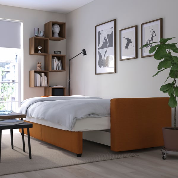 Modern bedroom with convertible bed, white covers, orange base, plant, bookshelves, & three art pieces.