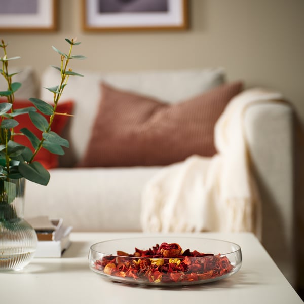 Glass bowl of dried flowers sits on a table, smaller than a plant in a vase. Sofa with pillows in background.