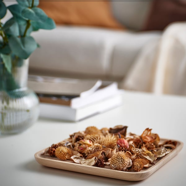 A small wooden plate of dried flowers sits on a table next to books and a glass vase.