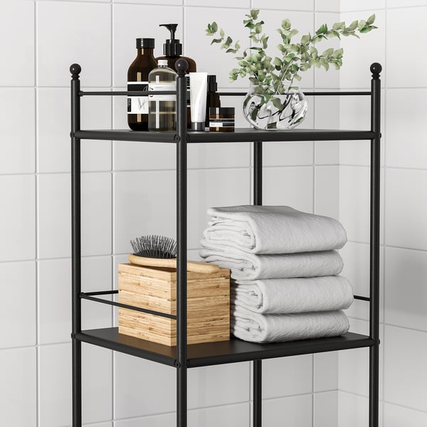 Black metal bathroom shelf holds stacks of white towels, bottles, and a plant in glass vase.