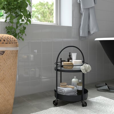 Small FRÖSJÖN black metal bathroom trolley with glass top, holding towels, brushes, and creams, next to a large wicker laundry basket and grey tiles.