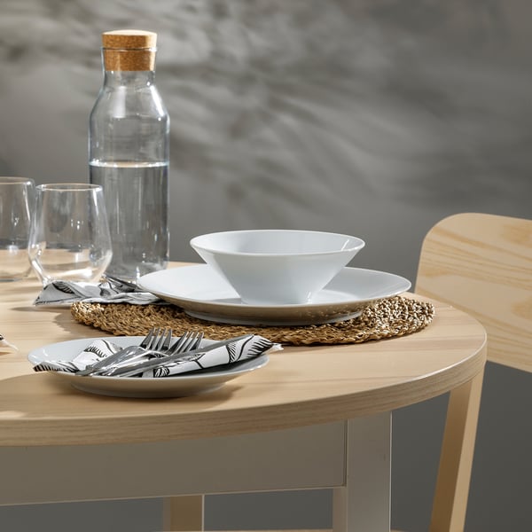 A light wooden circular dining table set for two with white plates, bowls, cutlery, and glasses.