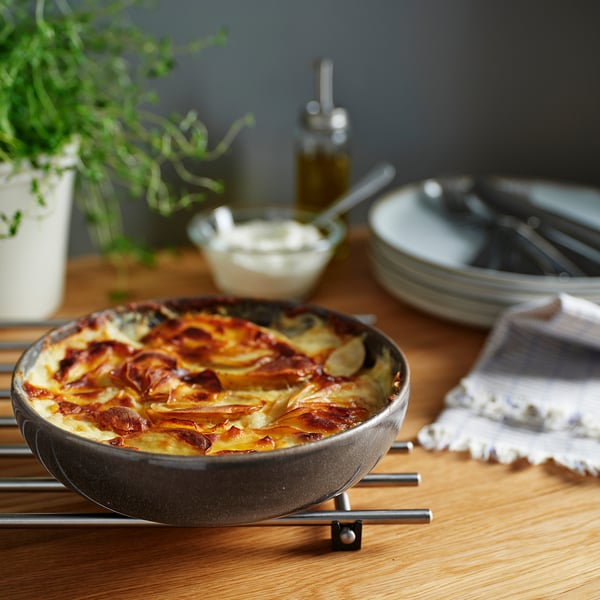 Grey oven dish on trivet with baked food, surrounded by plates, cutlery, and plant.