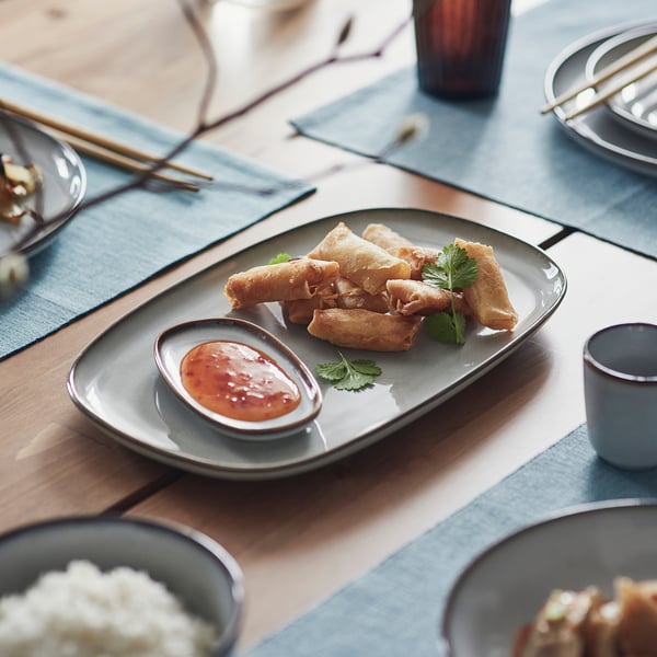 Plate of crispy spring rolls with dipping sauce on a stoneware platter, surrounded by bowls and chopsticks.