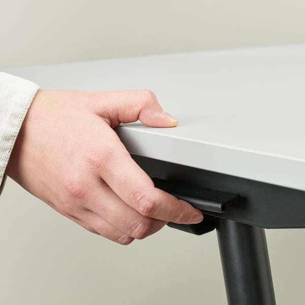 A hand adjusts a sit/stand desk, showing height adjustment feature. The desk has a white top, black base, and rubber feet for stability.