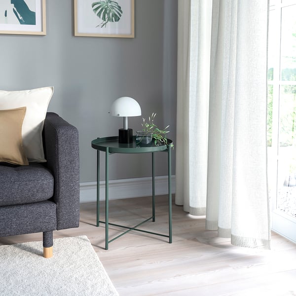 Small dark green side table with round tray top next to grey couch under white lamp.