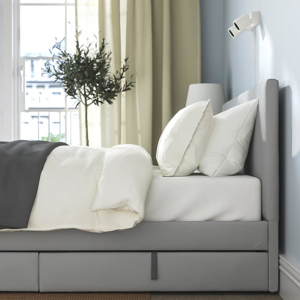 Modern bed with upholstered headboard and storage boxes, one visible, pull handles tucked; cream pillows, grey cover, neatly made.