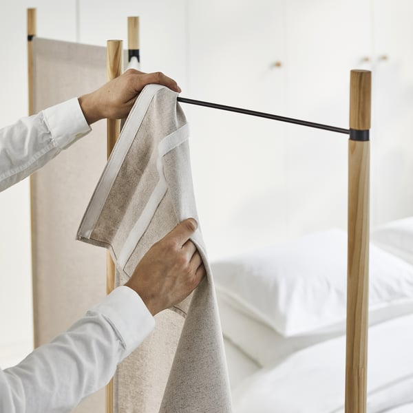 A person folds a beige GLAMBERGET foldable room divider, showcasing its lightweight and portable design.
