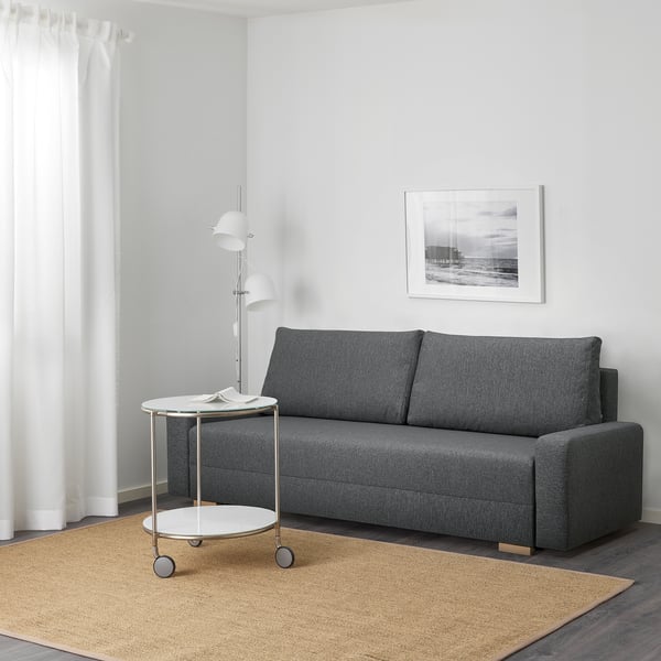 A modern living room a grey GRÄLVIKEN sofa-bed with a side table and lamp. The sofa-bed converts into a bed and has storage beneath the seat.