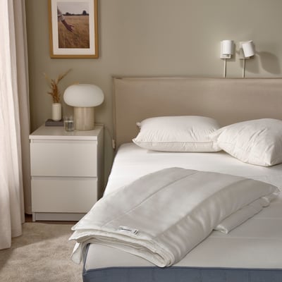 Modern bedroom with white bed, cream headboard, bedside table, lamp, vase, picture, curtains.