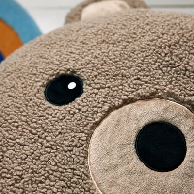 Adorable brown bear pillow, made from recycled polyester, cylindrical shape with friendly face detail.