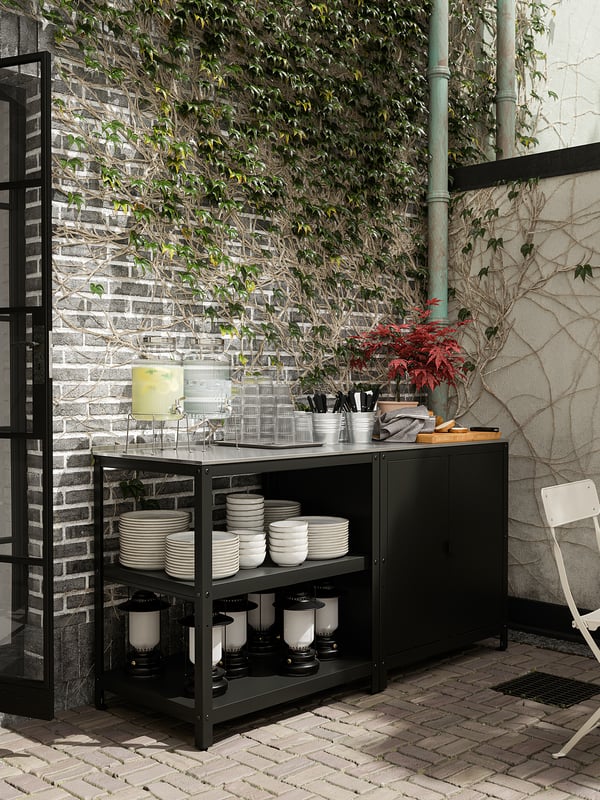 Black metal kitchen island with stainless top, white plates, and lanterns against brick and greenery.