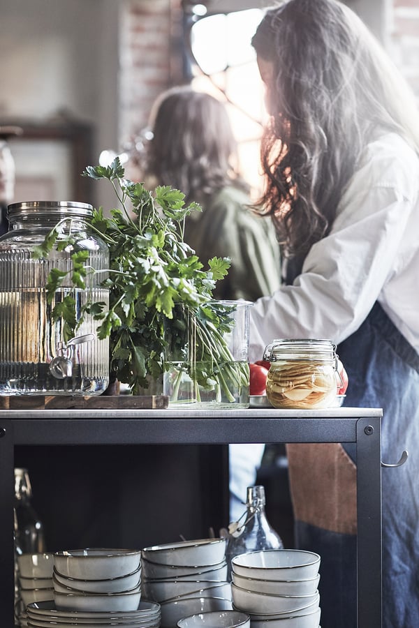 Person preparing food near a kitchen cart with glassware, herbs, and bowls. Sunlight streams in, highlighting the setup.