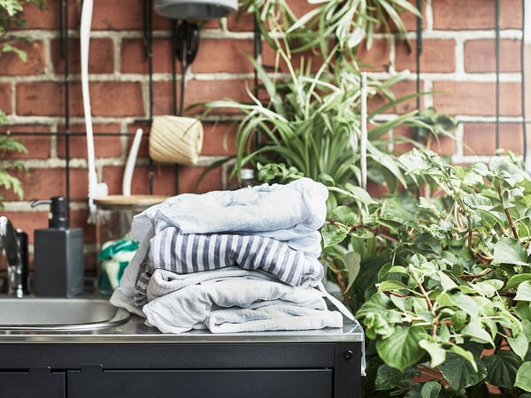 Stack of folded light blue/gray towels on black outdoor sink. Greenery and brick wall background.