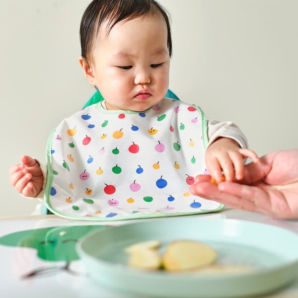 Baby wearing colourful bib with pockets, sits in high chair eating. The bib catches food, keeping clothes clean.