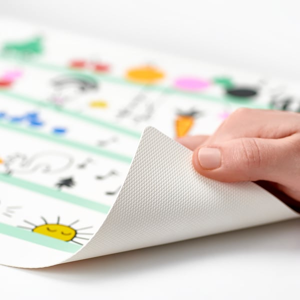 Hand peels back corner of colourful GRÖNFINK place mat, revealing grip pattern, designed for kids, protecting tables from spills.