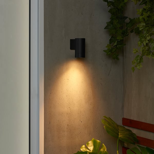 Small black GRÖNSPRÖT outdoor light, modern design, mounted high on wall, casting warm glow downward, surrounded by lush green plants.