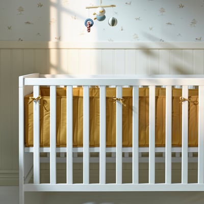 Image shows a baby crib with a padded bumper. The bumper, secured by ribbons, softens the hard slats and ensures safety.