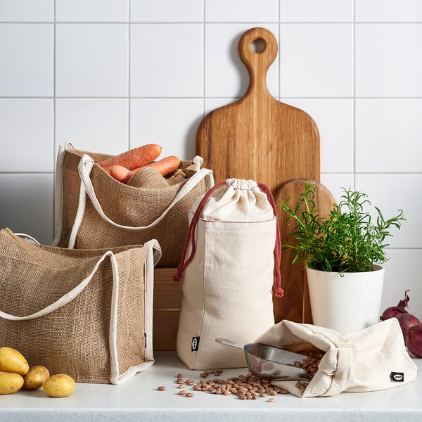 Natural hessian bags with white trim store root vegetables, beans. Wooden chopping boards, potted herb, red onion nearby.