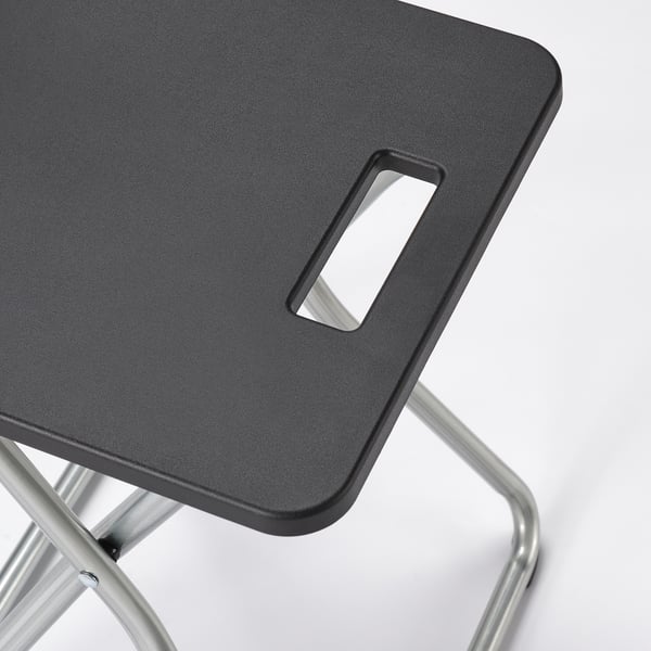 A modern, foldable stool with a black plastic seat and sturdy metal legs. A cutout handle is present for easy carrying.