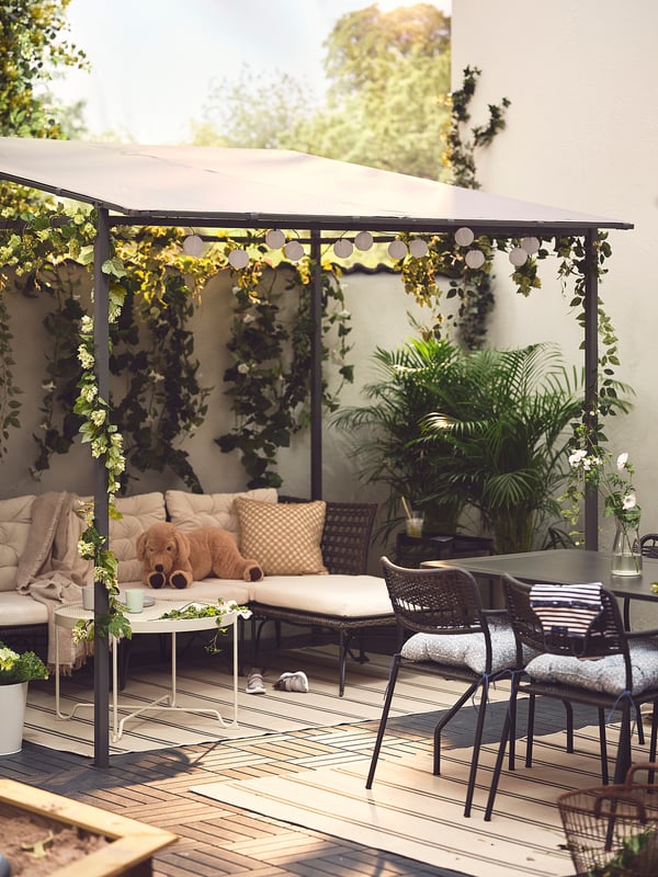 Outdoor seating area with beige GUNNÖN pergola, cushioned sofa, wicker chairs, hanging plants, and string lights, creating cosy, shaded patio space.