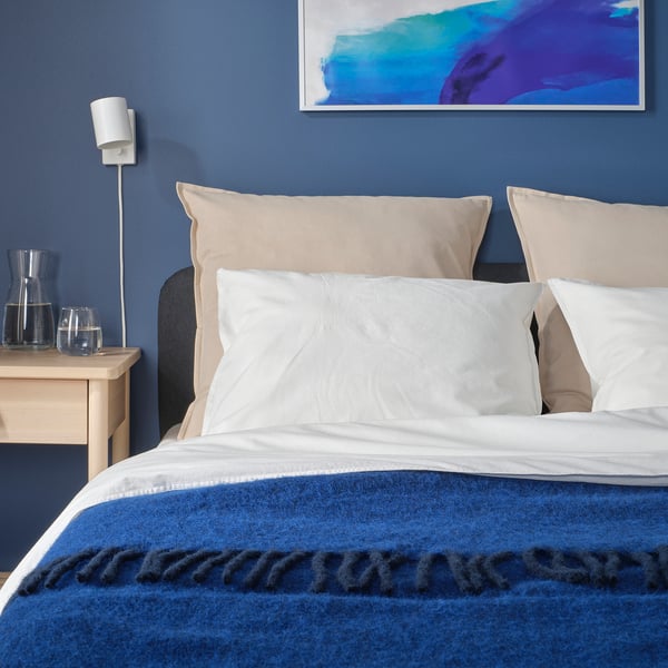 Modern bedroom with large bed, blue wall, and abstract art. GURLI pillow, light beige, covers visible. Two glasses beside bed.