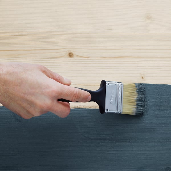 Paintbrush applying black paint on light wooden plank.