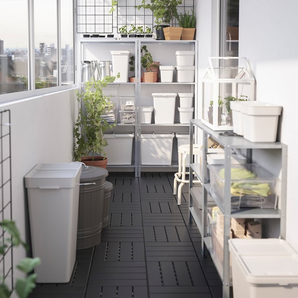 Balcony garden with grey HÅLLBAR bins, white shelves, potted plants, and city view.