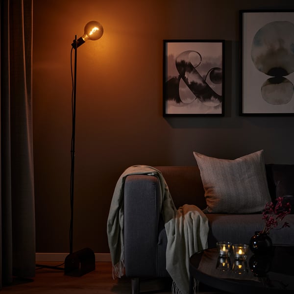 Cosy corner with HÅRSLINGA black floor lamp, grey sofa, cushions, blankets, and lit candles on table with art on wall.