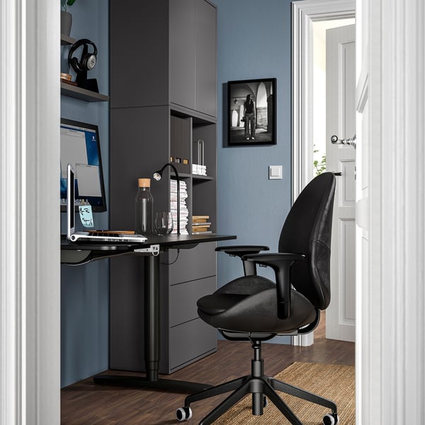 Home office with dark desk, ergonomic black leather chair, computer, shelves with books, and a framed photo on blue wall.