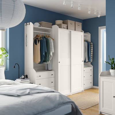 Modern blue bedroom with white HAUGA wardrobes, clothes, and plants.