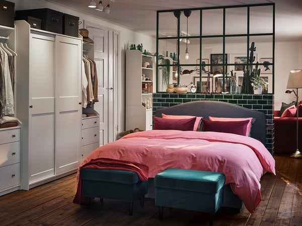 Modern bedroom HAUGA wardrobes in white, pink bed linens, blue ottomans, and glass-divided bed area.