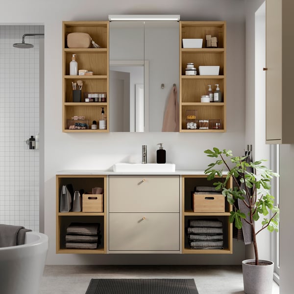 Modern bathroom vanity in beige, HAVBÄCK, with wood shelves, ceramic sink, and mirror. Plant, towels, and toiletries visible. Wall-mounted, spacious.