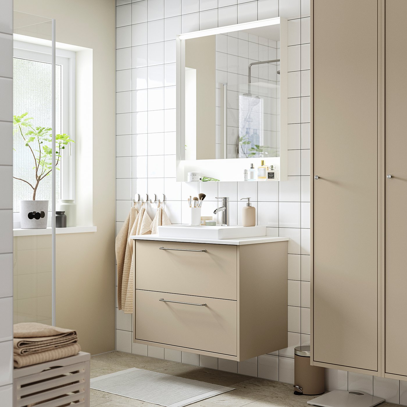 Modern bathroom with HAVBÄCK beige vanity. White counter, sink, matte finish. Mirrored cabinet, beige towels, and plants.
