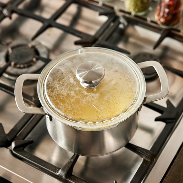 A saucepan with a glass lid simmers on a stove. The lid has a strainer and a handle.