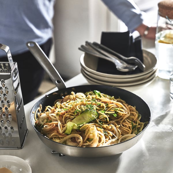 A person grates food with a box grater beside a pan of cooked noodles and greens in a frying pan.