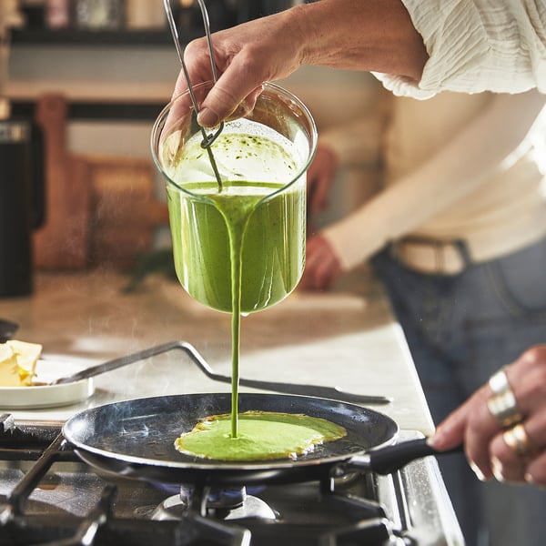 A person pours green batter from a glass into a non-stick frying pan on a stove, creating a flat, green pancake.