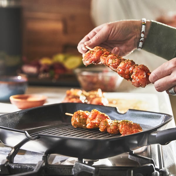 A hand places meat skewers onto a grill pan on a stovetop. The pan a non-stick ceramic coating, ensuring easy cooking and cleanup.