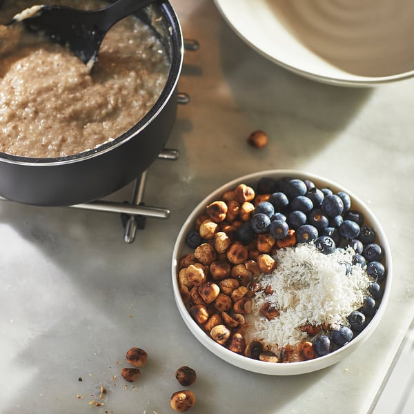 A saucepan on a stove, next to a bowl of oats topped with berries, hazelnuts, and cheese.