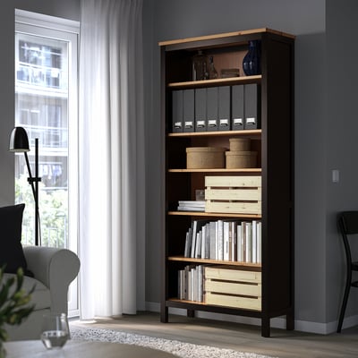 Dark brown HEMNES bookcase stands tall in corner, filled with books and storage bins, beside grey couch and lamp.