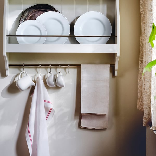 A kitchen shelf with white dishes and hanging towels. Four mugs hang from hooks and two towels hang from the shelf.