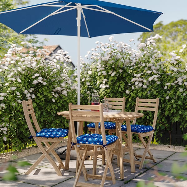 Outdoor patio set with blue parasol and cushioned chairs, surrounded by blooming bushes, ideal for summer relaxation.