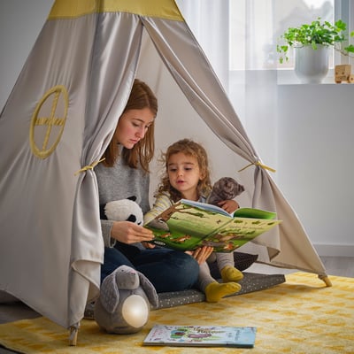 Young girl reads to toddler inside beige and yellow tent. Stuffed animals join them.