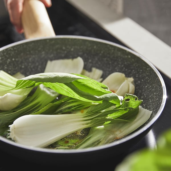 Cooking black pan with green vegetable, likely pak choi, ensuring even heat and easy cleaning.