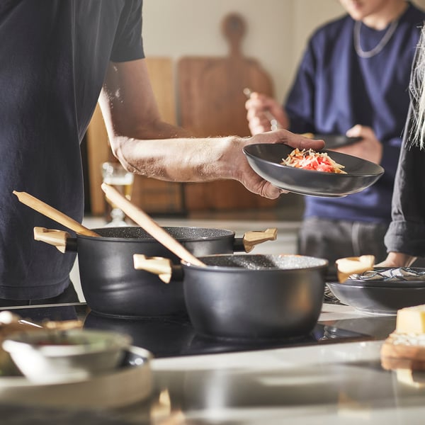 Chefs prepare food in modern kitchen with non-stick pots. One serves dish, another cooks on stove.