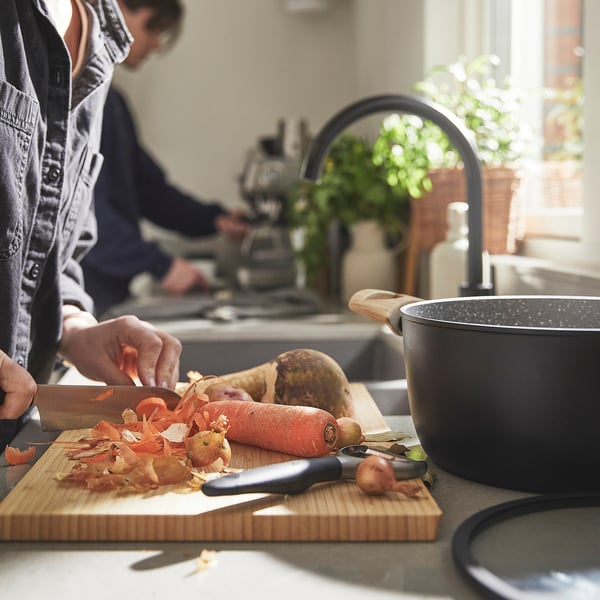 A person is peeling vegetables on a cutting board. A saucepan with a lid sits nearby, ready for cooking.