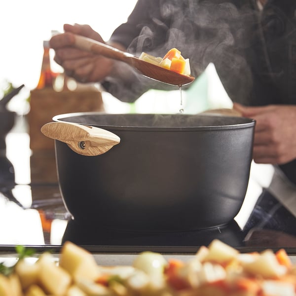 Person ladling steaming food from a black saucepan. The saucepan has wooden handles and a vented lid.