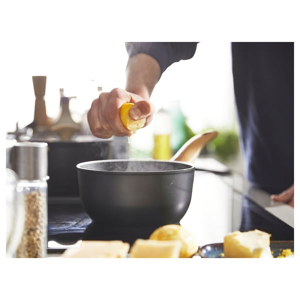 Hands squeeze lemon over a black saucepan on a stove. A cutting board with lemons and butter nearby. Kitchen utensils visible.