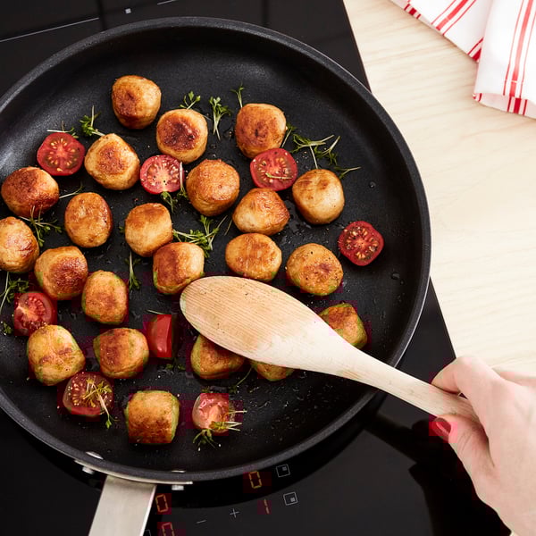 Black skillet with golden-brown fried foods, halved tomatoes, and green herbs. Hand holds wooden spoon in the pan.