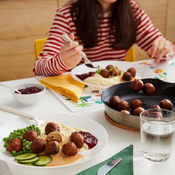 A person in a striped shirt eating from a plate of meatballs, mashed potatoes, peas, and cucumbers on a white table with a glass of water.